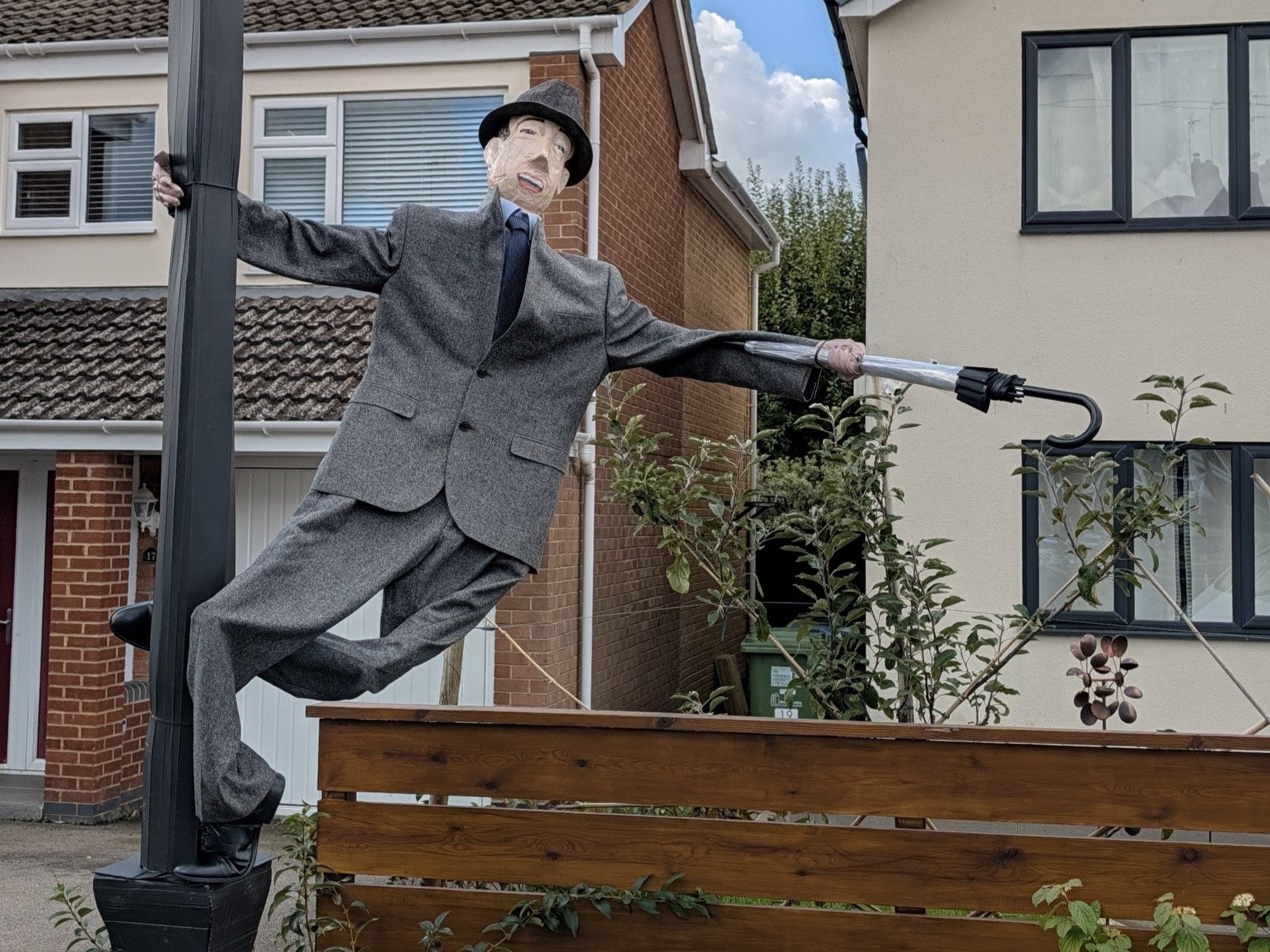 Scarecrow dressed as Gene Kelly stands on a lamppost holding a closed umbrella, in the iconic pose from Singin' in the Rain