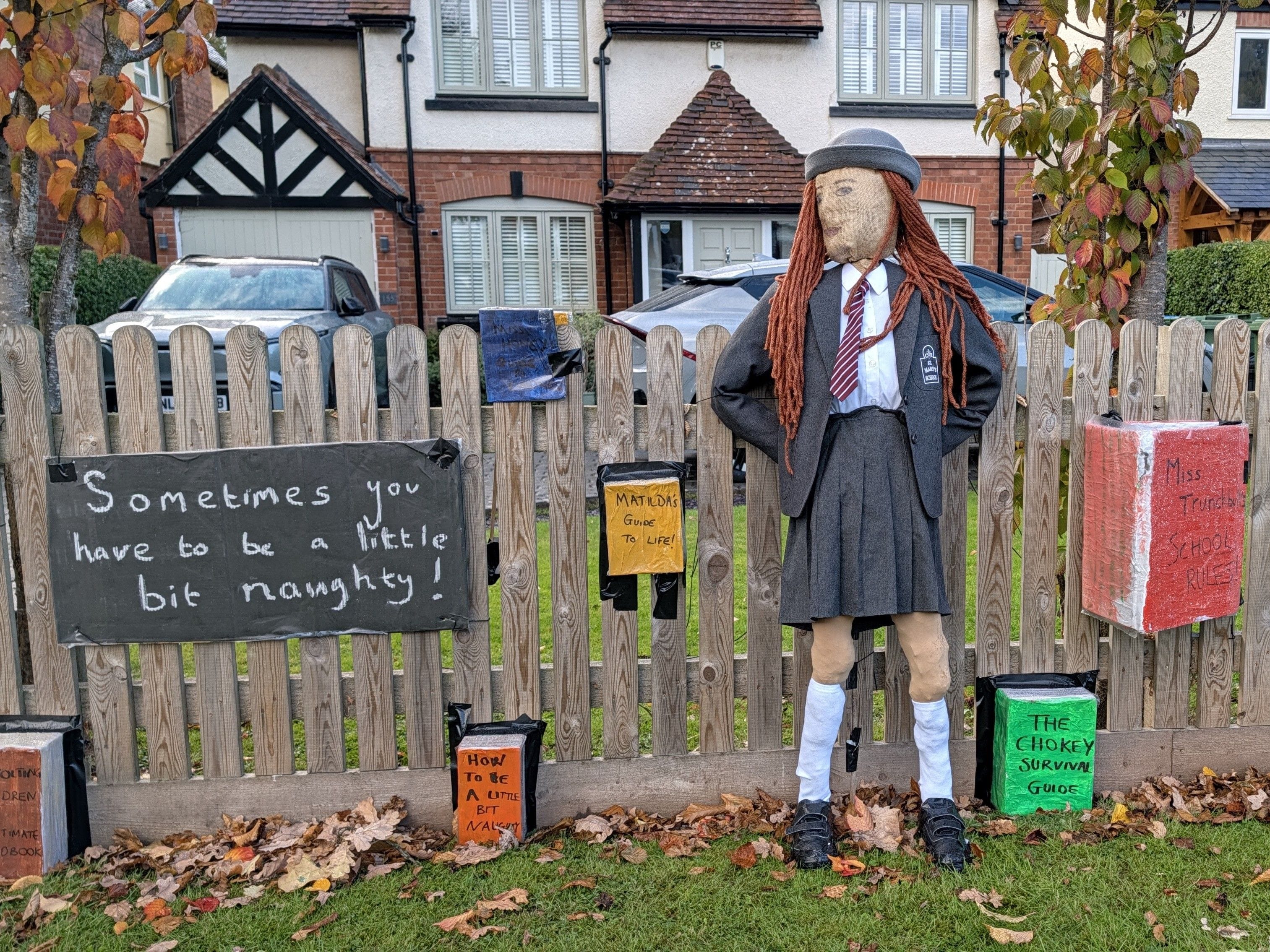 Scarecrow dressed as Matilda stands in front of a garden fence, surrounded by books.