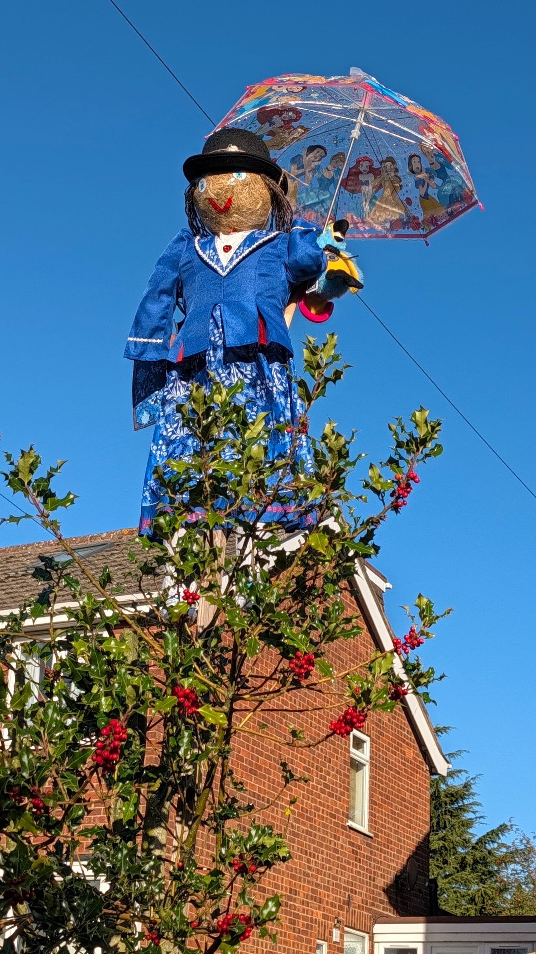 Scarecrow dressed as Mary Poppins  and holding a child's umbrella stands on top of a tree to look like she's flying.