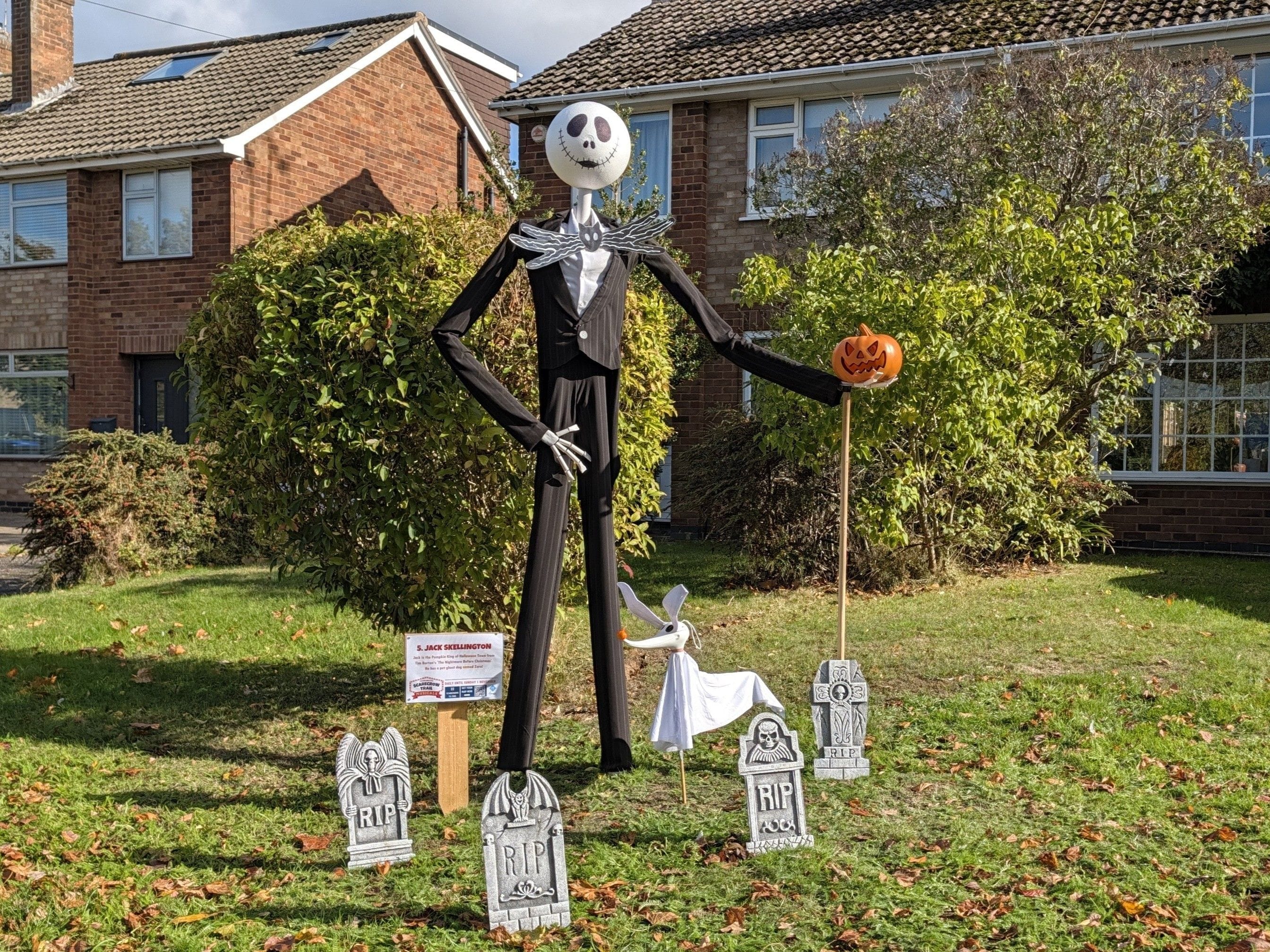 Tall scarecrow Jack Skellington holds a small pumpkin and poses in a front garden with his dog Zero. They are surrounded by small headstones.